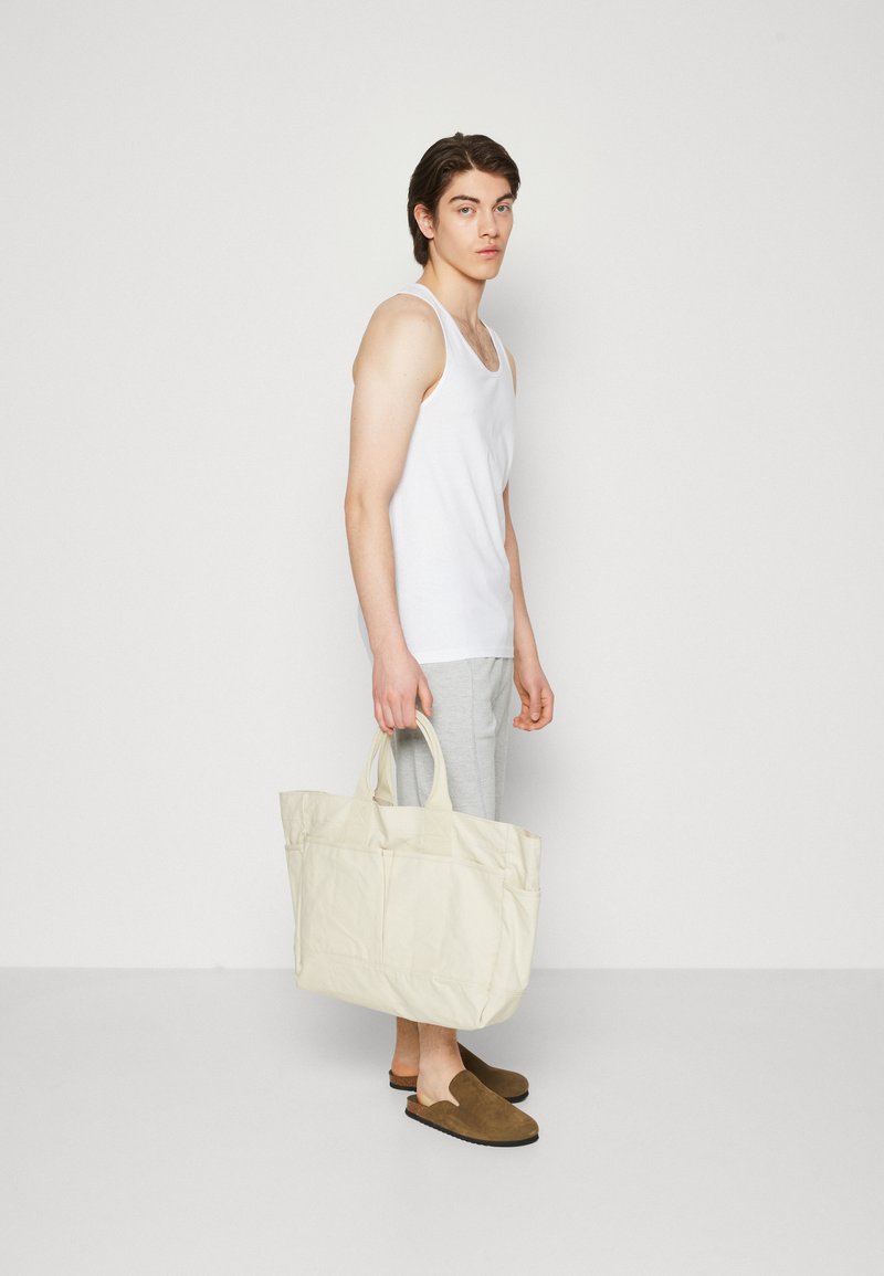 Canvas-Tragetasche in hellem Creme mit zwei oberen Griffen, die eine strukturierte Form und Außentaschen aufweist, kombiniert mit grauen Jogginghosen und braunen Slippern.