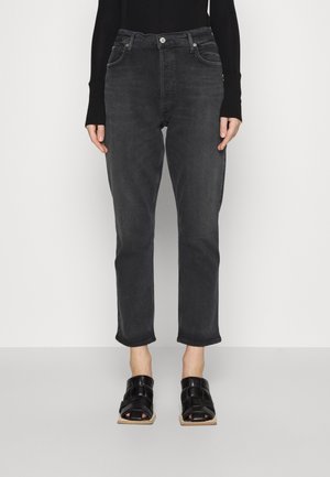 Personne portant un jean noir taille haute, un haut noir à manches longues et des sandales noires à bout carré avec des brides tissées, debout devant un fond uni.