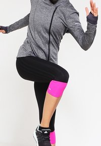 Osoba w szarej bluzie z zamkiem i czarnych legginsach z różowymi ochraniaczami na kolana i kostki, podnosząca jedną nogę, w czarnych trampkach Adidas.