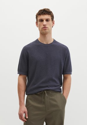 Młody mężczyzna stoi z rękami w kieszeniach, ubrany w granatowy, krótki sweter z dzianiny i oliwkowe spodnie ze sznurkiem, jednolite białe tło.