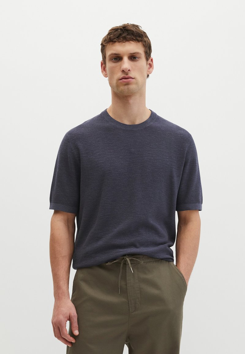 Junger Mann steht mit den Händen in den Taschen, trägt ein marineblaues kurzärmeliges Strickshirt und olivgrüne Hose mit Kordelzug, einfarbiger weißer Hintergrund.