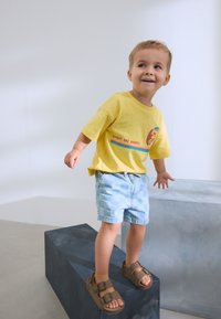 Maglietta gialla a maniche corte con la scritta "RAGGI DI SOLE E ONDE", pantaloncini a quadretti azzurri chiari e sandali marroni con due fibbie.