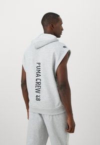 Siva brezrokavnik s kapuco, na hrbtu z napisom "PUMA CREW '48" v debelem črnem tisku. Mehka tkanina in sproščen kroj.