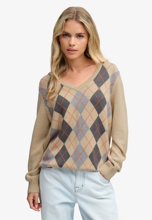 Lyshåret kvinde iført en beige argyle sweater med grå og sorte diamantmønstre og lyseblå jeans, står med den ene hånd i lommen.