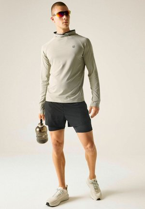 Hombre con camisa gris de manga larga, pantalones cortos negros, zapatillas blancas y gafas de sol sostiene una botella de agua oscura sobre un fondo liso.