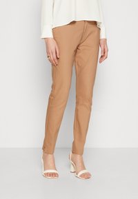 Žena v hnědých slim kalhotách, bílé blůze s dlouhým rukávem, bílých podpatcích a zlatém prstenu na levé ruce.