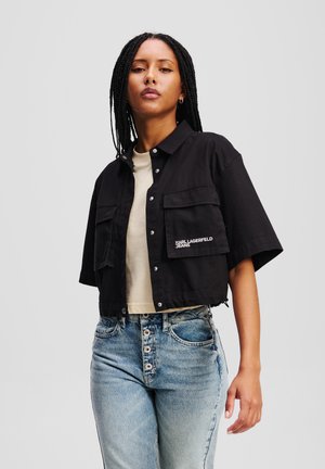Maglietta nera corta con maniche corte, dotata di due tasche frontali e un logo bianco. Indossata sopra un top beige chiaro e abbinata a jeans blu a vita alta.