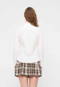 Λευκό πουκάμισο με μακριά μανίκια και καμπυλωτό τελείωμα, συνδυασμένο με καφέ πλισέ φούστα σε καρό σχέδιο. Το ύφασμα φαίνεται ελαφρύ και απαλό.