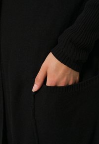 Mano parzialmente dentro alla tasca di un capo in maglia nera con polsini a costine.