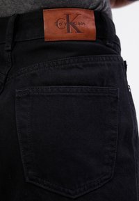 I pantaloni in denim neri presentano una caratteristica etichetta in pelle con la scritta "Calvin Klein" e un logo impressa. Il tessuto è resistente e ha una texture liscia.