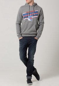 Sudadera con capucha gris que presenta un gráfico de "New York Rangers" en rojo y azul, combinada con jeans de mezclilla oscura y zapatillas negras.