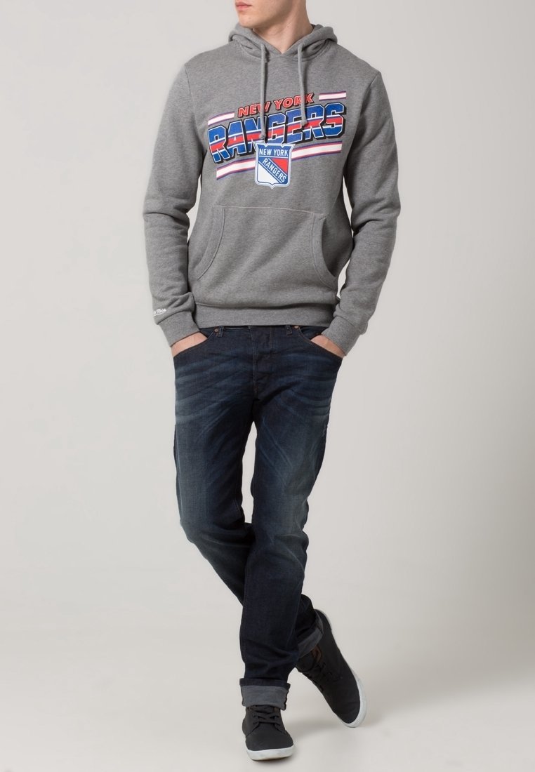 Sudadera con capucha gris que presenta un gráfico de "New York Rangers" en rojo y azul, combinada con jeans de mezclilla oscura y zapatillas negras.