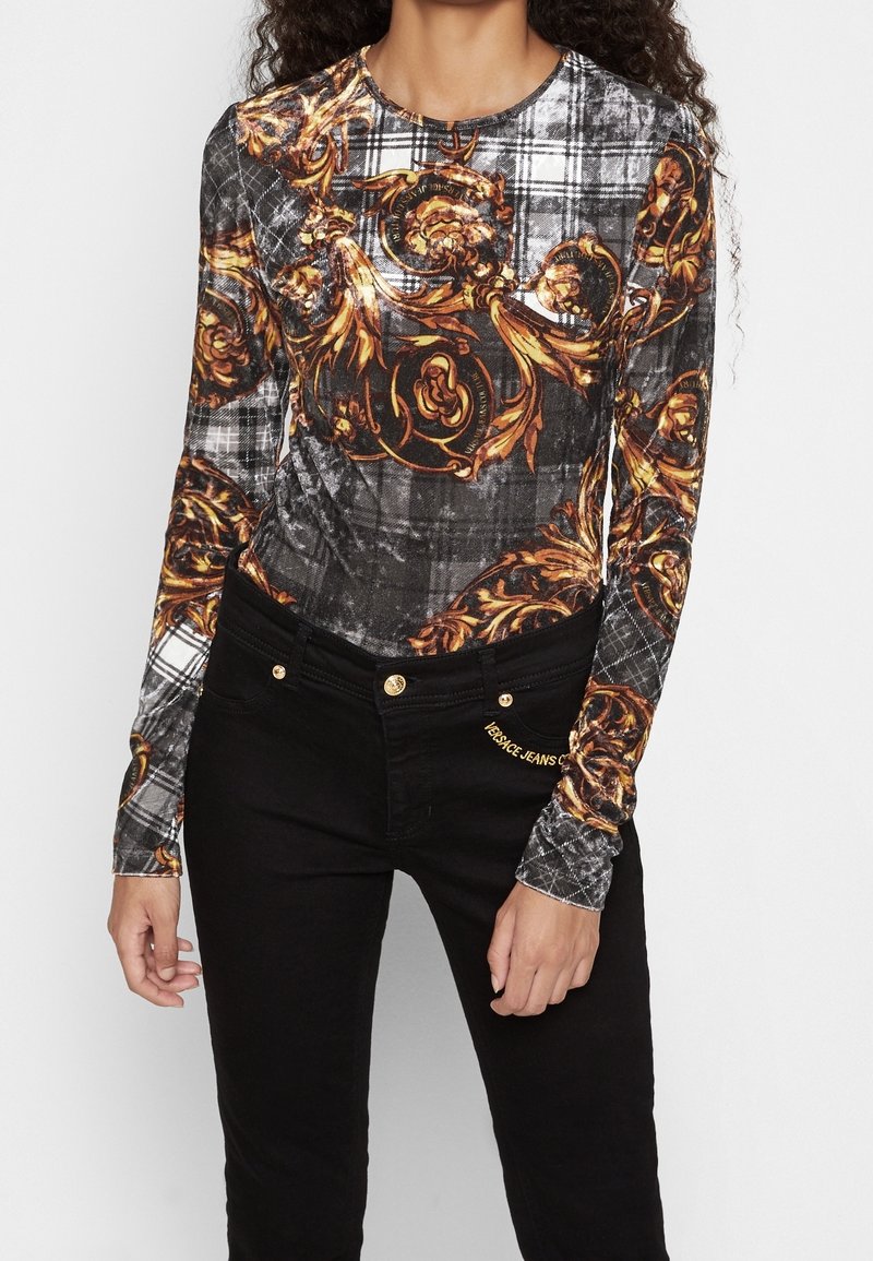 Långärmad skjorta i svart och grått mönster med guldblommor och barockmönster, kombinerad med svarta jeans som har guldiga detaljer.