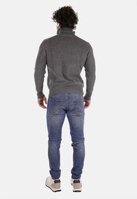 Maglione grigio a coste con colletto alto, abbinato a jeans in denim blu. Il modello indossa sneakers chiare, visto da dietro.