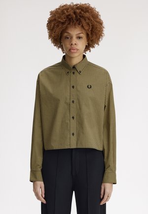 Woman with curly hair wears a cropped, long-sleeve checkered shirt with black buttons and black high-waisted pleated pants.