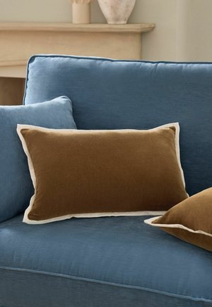 Blaues Stoffsofa mit zwei braunen rechteckigen Kissen mit hellbeigen Kanten, in einem neutral gehaltenen Raum mit Kaminsims und dekorativer Vase.