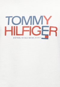 Λευκό βαμβακερό T-shirt με έντονο κόκκινο και μπλε κείμενο "TOMMY HILFIGER ESTABLISHED MCMLXXXV" σε μοντέρνα, μπλοκ γραμματοσειρά.