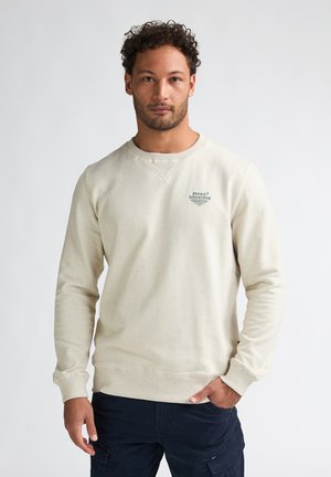 Man met krullend haar draagt een lichtbeige trui met lange mouwen en een klein Petrol Industries-logo op de borst, staande tegen een effen achtergrond.