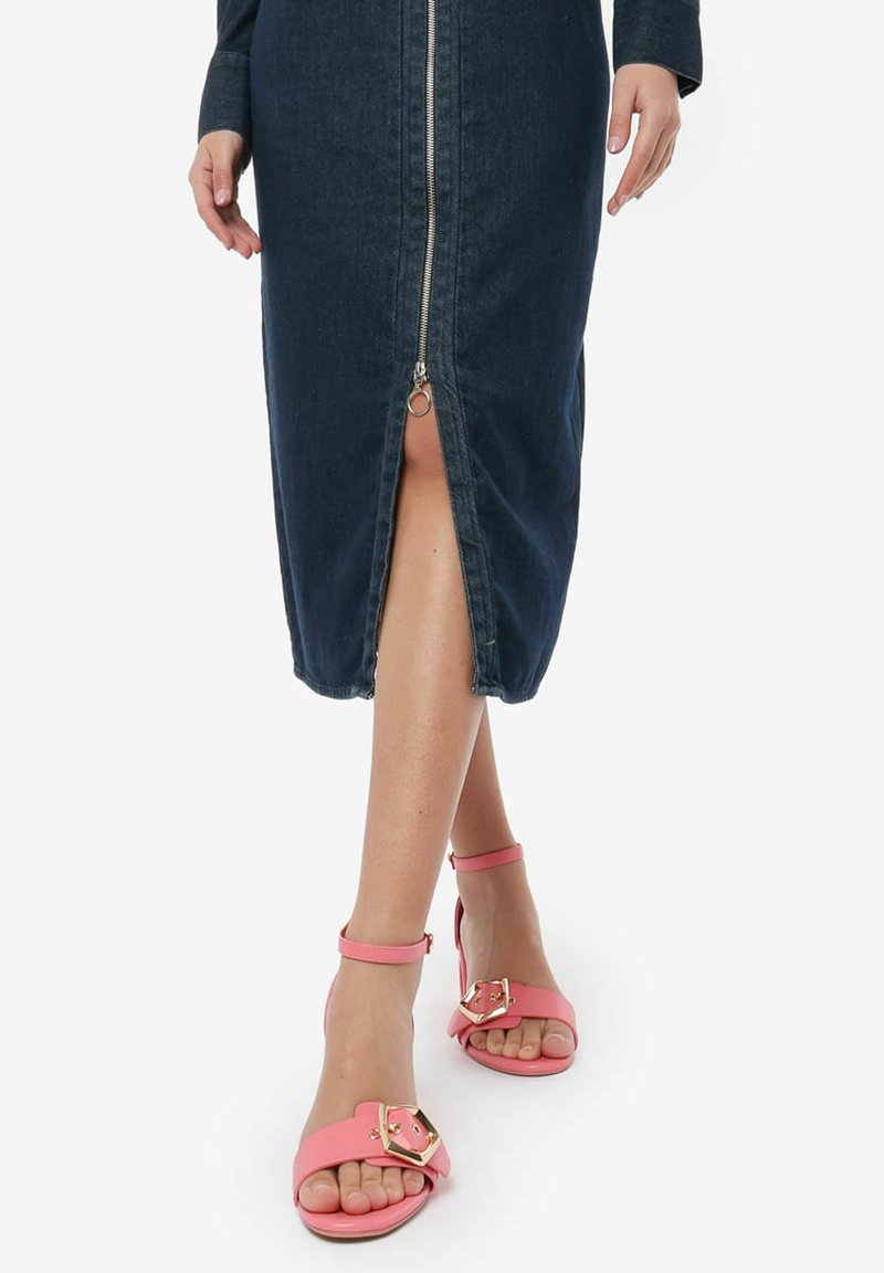 Donkerdenim midi-jurk met een ritssluiting aan de voorkant en een split. Roze sandalen met een brede strap en een gouden decoratieve gesp maken de look compleet.