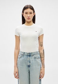 Jauna sieviete ar gariem tumšiem matiem valkā piegulošu baltu T-kreklu un gaiši zilas augsta vidukļa džinsas, uz kreisās rokas redzami tetovējumi.