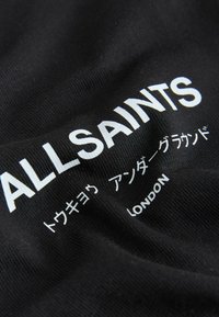 Tela negra con texto blanco "ALLSAINTS" y caracteres japoneses más pequeños, junto con la palabra "LONDON" impresa.