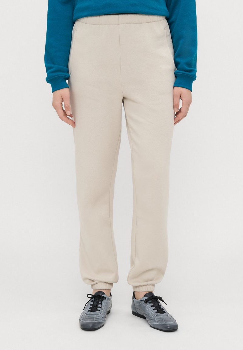 Beige sweatpants met elastische tailleband en boorden. Gemaakt van zacht materiaal, met een gladde textuur en een ontspannen pasvorm. Gekozen in combinatie met grijze sneakers.