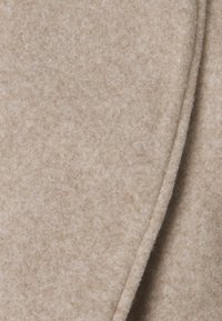 Tessuto di lana di un beige chiaro con una texture morbida, simile al feltro, e una superficie leggermente rialzata. Il materiale presenta una finitura liscia con variazioni tonali.