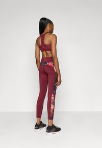 Burgundi színű sport crop top, kulcslyuk hátkivágással, burgundi leggingsszel párosítva, amelyek virágmintás díszítéssel és fekete oldalpanellel rendelkeznek. Fekete sportcipők.