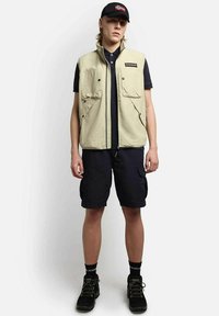 Chaleco sin mangas beige con dos bolsillos frontales, camiseta tipo polo negra, pantalones cortos cargo oscuros, botas de senderismo negras y una gorra negra con un diseño de bandera.