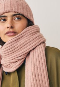 Lichtroze geribbelde gebreide muts en bijpassende sjaal gedragen over een olijfgroene jas. De sjaal heeft een dikke, zachte textuur.