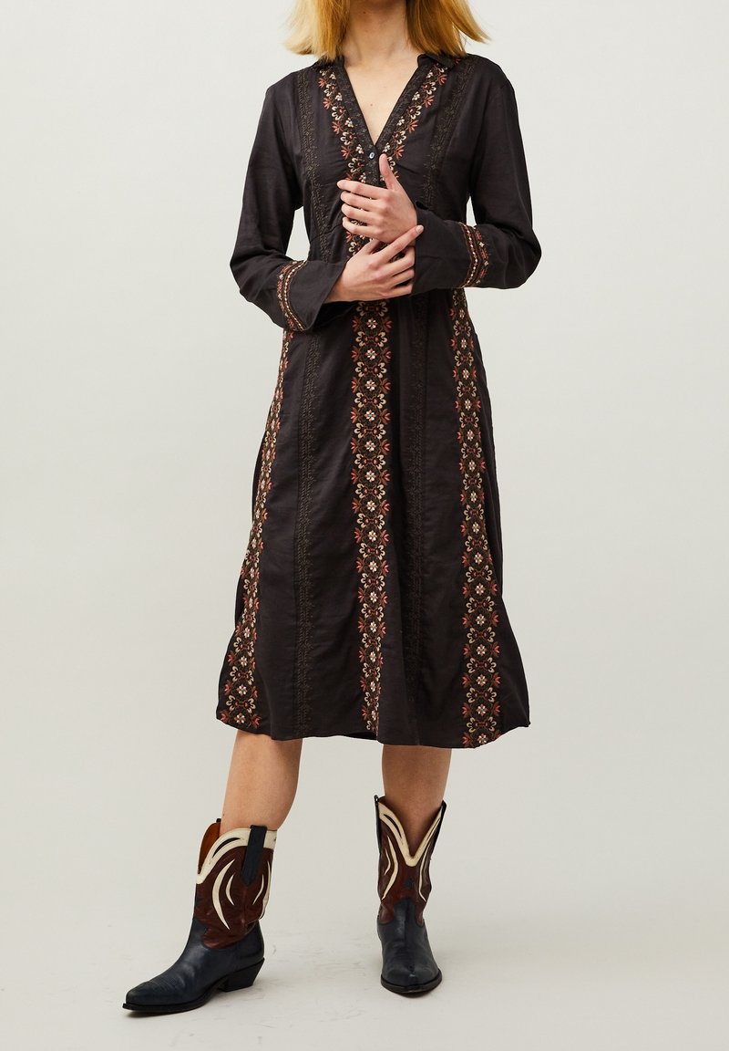 Femme portant une robe midi noire avec une broderie florale verticale et des bottes de cowboy noires et marron de style western.