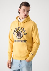 Haut à capuche jaune en coton, avec un design graphique portant "GANT 1949" en bleu foncé. Comprend une poche avant et un cordon de serrage.