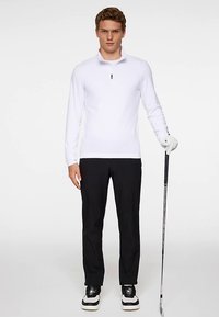 Vit golftröja med långa ärmar och halvlång dragkedja vid kragen, kombinerad med svarta byxor och vita sneakers, håller i en golfklubba.