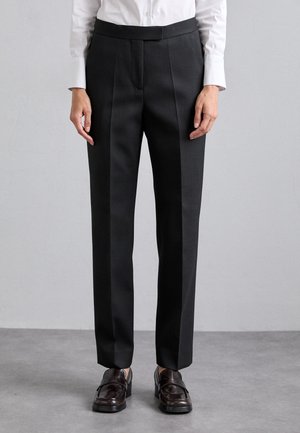 Person trägt schwarze maßgeschneiderte Hose, weißes Hemd mit Knöpfen und dunkelbraune Leder-Loafer, steht auf grauem Boden vor heller Wand.