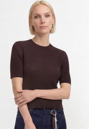 Lyshåret kvinde iført en mørk ribbet kortærmet top og mørke jeans med reb-bælte, stående med den ene arm på tværs af kroppen.
