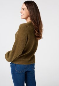 Olijfgroene donzige trui met een relaxte pasvorm, ronde hals en lange mouwen, gecombineerd met strakke blauwe jeans. Gezichtsdetail met textuur zichtbaar.