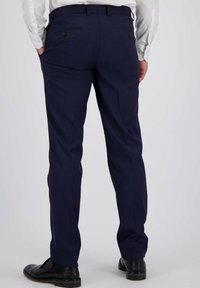 Man in een donkerblauwe formele broek, wit overhemd en zwarte nette schoenen, staande met handen in de zakken, gezien van achteren.