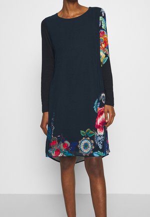 Kvinde iført navyblå kjole med lange sorte ærmer og farverige blomsterprint på nederste og højre side mod ensfarvet lys baggrund.