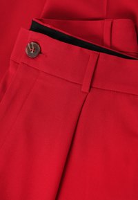 Pantalon rouge sur mesure en tissu tissé. Présente un devant plat, des poches latérales et un détail de bouton marron. Texture lisse avec une finition nette.