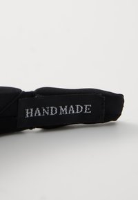 Musta käsintehty tekstiilituote, jossa on valkoinen "HAND MADE" -merkki, näkyvää ompelua ja pehmeä, sileä pinta.