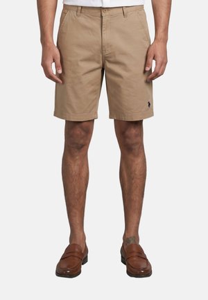 Man draagt beige knielange shorts en bruine leren loafers, staat met ontspannen armen langs het lichaam tegen een effen achtergrond.