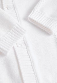 Cardigan en maille blanche douce avec poignets côtelés et détails de boutons sur un fond de tissu texturé.