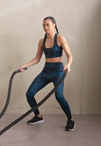 Ropa de fitness en azul oscuro con un patrón de hojas; top corto y leggins de cintura alta. Teniéndo que esforzarse contra cuerdas gruesas, usa zapatos negros.