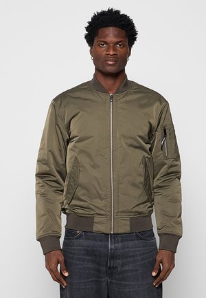 Mann trägt olivgrüne Bomberjacke mit Frontreißverschluss und dunkle Jeans, steht vor einfachem hellem Hintergrund.