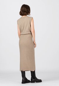 Stickad, ärmlös beige klänning med ribbad textur, figurnära midja och lång fåll. Bärs med svarta ankellånga stövlar för kontrast.