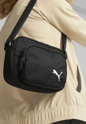 Sac bandoulière Puma noir avec logo blanc porté sur une veste beige, montrant la poche avant zippée et la sangle réglable.