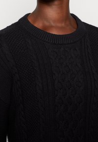 Mustat neulotut neuleet, joissa on pyöreä pääntie, tekstuuriset palmikkoneulekuviot sekä ribbaantuneet hihansuut ja helma. Lähikuvassa yksityiskohtia.