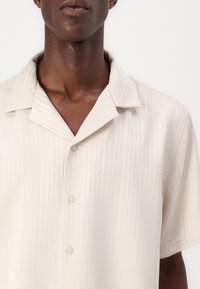 Kortermet, lys beige knappeopp skjorte med en subtil vertikal stripe tekstur og en klassisk krage design.