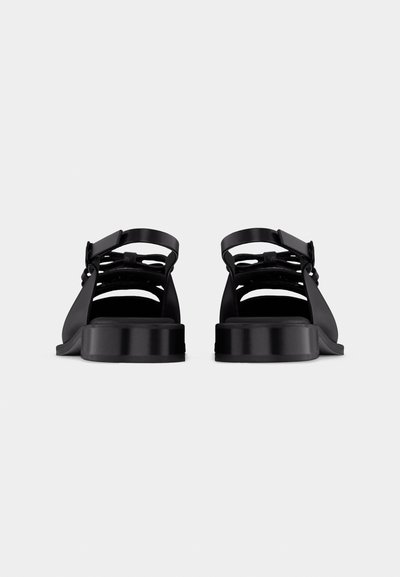 Ζευγάρι μαύρων σανδαλιών με φαρδιές λουρίδες και χαμηλά τακούνια σε σχήμα μπλοκ, εμφανιζόμενα από πίσω σε απλό λευκό φόντο.