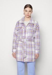 Manteau à carreaux violet et blanc avec un col, des poches avant, une taille ceinturée et une texture duveteuse. Porté par-dessus un pull léger et un pantalon bleu.